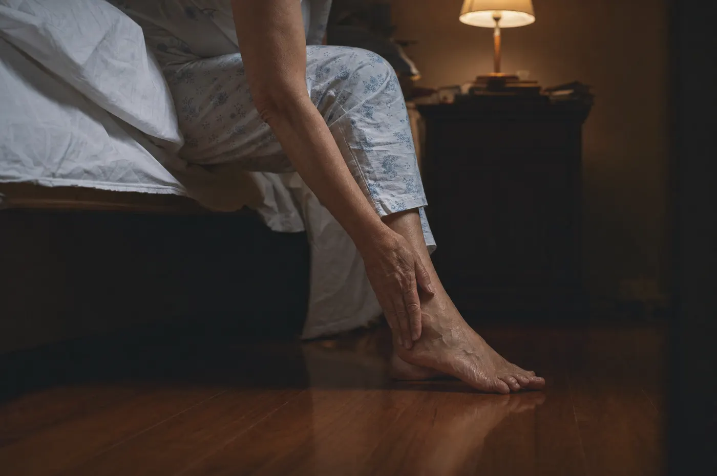 Persona sentada en la orilla de la cama por la noche, sobándose el pie